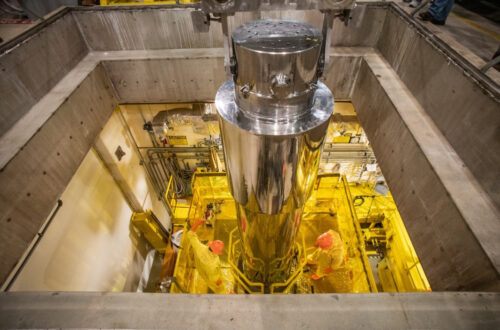 To prepare for extraction, personnel at the tritium extraction facility receive a cask of irradiated rods from the Tennessee Valley Authority. The cask can be seen from the top viewed through a room built in concrete. It has a shining steel surface and cylindrical shape. It appears to be at least three times as high as the operator. The person wears a yellow protective suit, orange helmet and gloves.